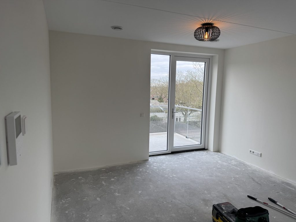 Lege nieuwbouwkamer met lichte wanden, een betonvloer en openslaande deuren naar een balkon